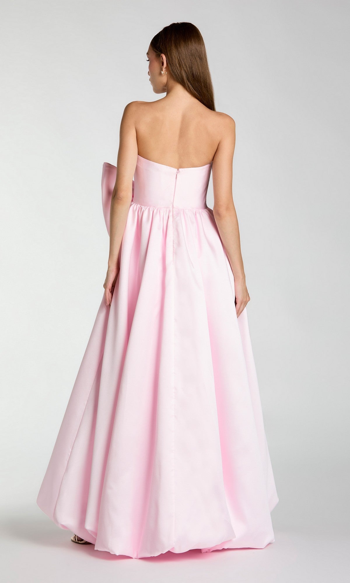 A woman with long straight hair stands with her back to the camera against a plain background, wearing the Mac Duggal 21245—a stunning, floor-length pastel pink formal dress with a strapless neckline and full skirt.